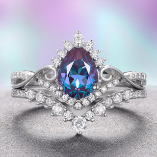 A two-piece ring set with a teardrop-shaped alexandrite centerpiece in 14K white gold, accompanied by a band with intricate leaf detailing. The ring is displayed against a gradient purple background.