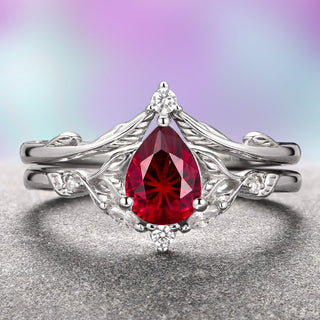 A two-piece gold ring set, featuring a teardrop-shaped ruby center stone with a feather design band, placed against a purple and gray background.
