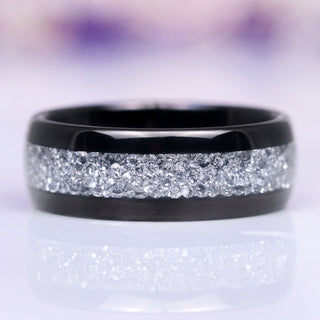 A polished black tungsten carbide wedding band with an 8 mm width, featuring a silver stone inlay creating a sparkle effect, displayed against a floral background.