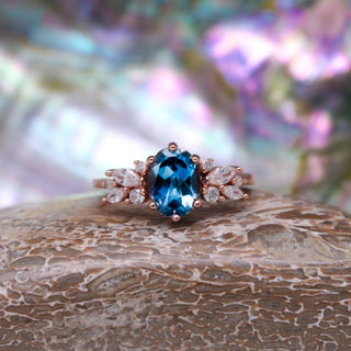 Front view of the Lily London Blue Topaz Ring in 14K Rose Gold Vermeil by Aquamarise on a textured stone, with mother-of-pearl shimmer behind