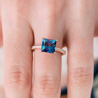 Rose gold ring with princess cut alexandrite center stone and diamond accents on hand over a light background 