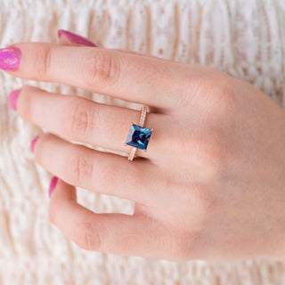 Rose gold ring with princess cut alexandrite center stone and diamond accents on hand over a light background 