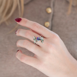 Silver leaf guard wedding band with emerald and diamond accents and oval mystic topaz engagement ring displayed in hand against warm brown backdrop
