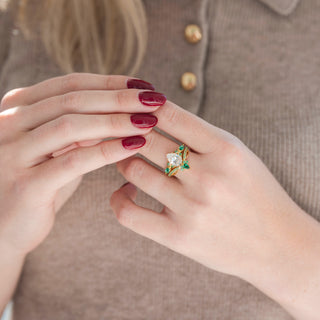 Yellow gold crown tiara-inspired wedding band and pear-shaped diamond engagement ring with emerald accents displayed in hand against warm brown backdrop
