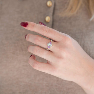Oval-shaped moissanite engagement ring with pink sapphire accent stones displayed in hand against warm brown backdrop