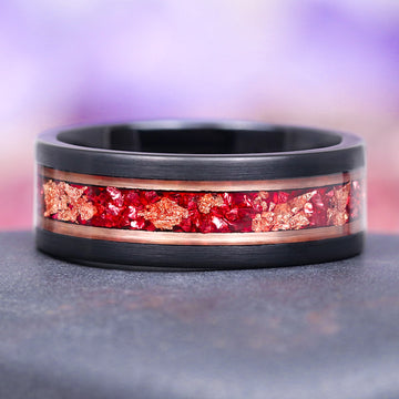 black tungsten carbide men's wedding band with ruby and rose gold inlay on a gray surface with a blurred purple background