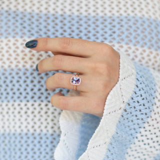 Hand wearing a rose gold ring with a purple gemstone against a textured light blue and white background