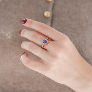 Rose gold oval-shaped alexandrite engagement ring with diamond accent stones and leaf motif wedding band displayed in hand against warm brown backdrop