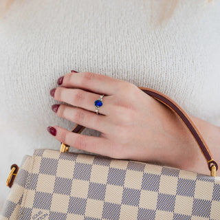 Oval Cut Blue Sapphire Engagement Ring in Sterling Silver by Aquamarise featuring a vibrant blue sapphire with delicate silver detailing, styled on a hand holding a checkered designer handbag.
