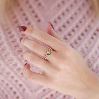 Rose gold leaf vine motif green moss agate engagement ring with diamond accent stones displayed in hand against warm pink backdrop