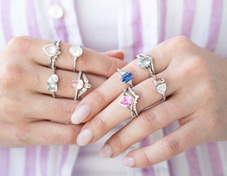 Aquamarise Two hands wearing multiple rings with various gemstones against a striped background