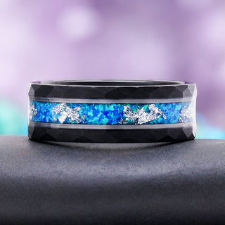 8mm Blue Fire Opal men's wedding band in hammered black tungsten carbide resting on a dark stone, showing the blue opal inlay and silver rails