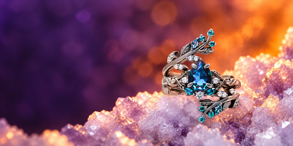 Pear-cut blue gemstone engagement ring with leaf and vine shoulders and teal accents in black rhodium, on amethyst crystals with purple and amber bokeh, Aquamarise banner