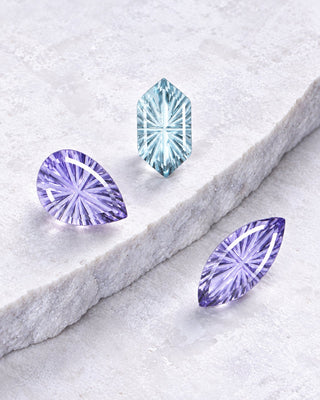 Three fantasy-cut loose gemstones on white marble: lavender pear, aqua hexagon and lavender marquise with starburst facets, Aquamarise banner