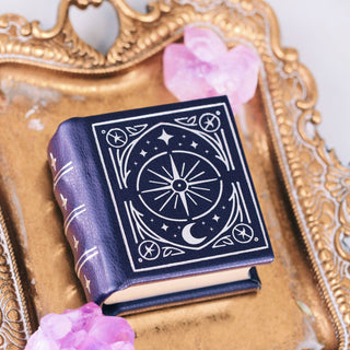 Small dark blue book with a celestial design on the cover resting on an ornate gold tray, surrounded by pink crystal clusters on a light surface.
