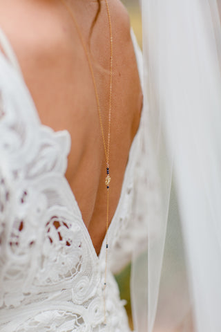 Delicate gold chain necklace with small dark beads draped along the back of a lace wedding dress, complemented by a soft white veil on the bride's shoulder.