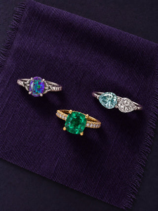 Three engagement rings on a frayed purple swatch: lavender opal and emerald fairy leaf set in white gold, cushion-cut emerald hidden halo ring in yellow gold and toi et moi blue moissanite ring in white gold, Aquamarise banner