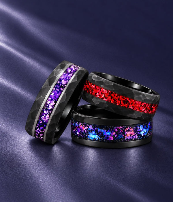 Three black men's wedding bands stacked on navy satin: hammered with purple amethyst inlay, hammered with crushed red inlay and polished with pink-blue galaxy inlay, Aquamarise banner