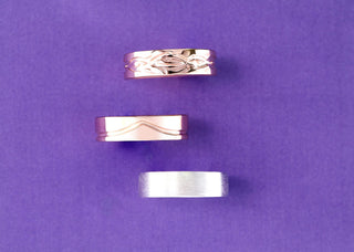 Three men's wedding bands on purple fabric: a scroll-engraved rose gold band, a wave-grooved rose gold band, and a brushed silver band.