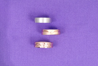 Three men's wedding bands on purple fabric: a brushed silver band, a rose gold band with a wave groove, and a polished rose gold band.
