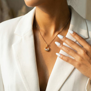Person wearing a gold necklace with a pendant, hand touching face, blurred background