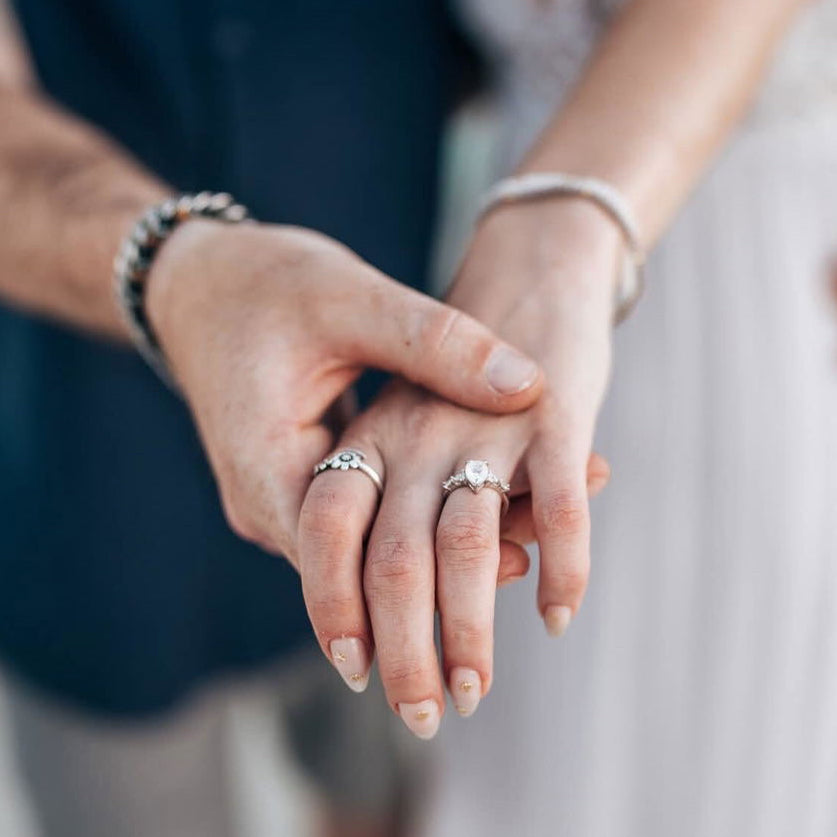 Customer photo of a couple holding hands outdoors, showing a pear-cut Aquamarise engagement ring and a matching men's band