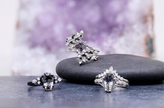 Two black rutilated quartz rings in sterling silver resting on dark slate and a grey pebble against an amethyst geode.