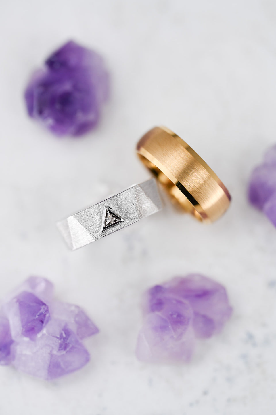Solid gold men's wedding bands, one yellow gold and one white gold with a triangle design, beside purple crystals