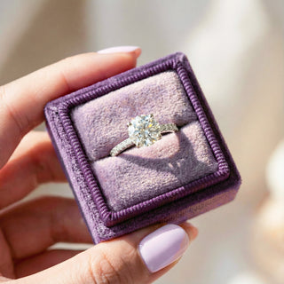Engagement ring in a purple box held by a hand with light pink nail polish.