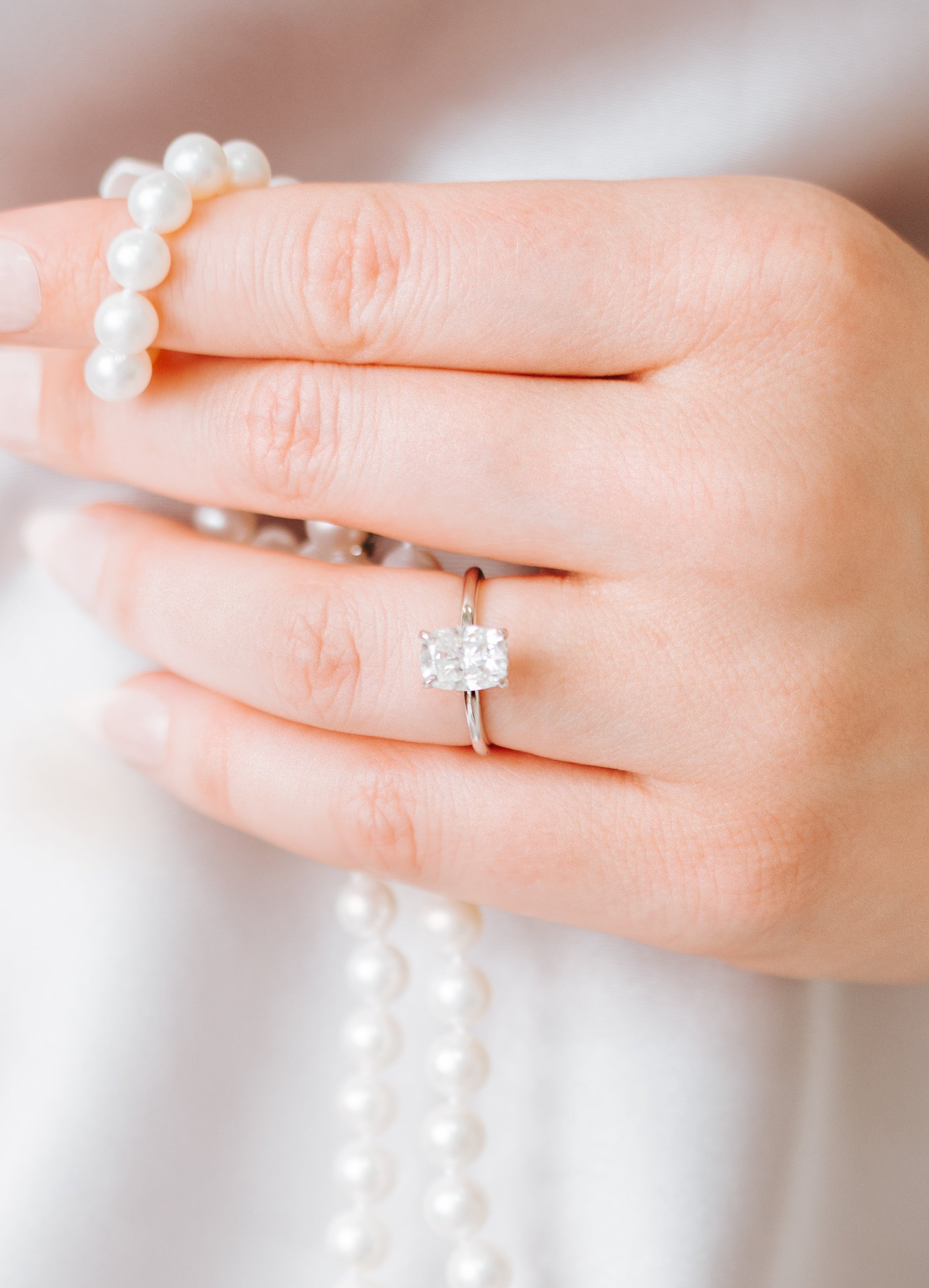 Elongated cushion cut solitaire engagement ring on a thin white metal band, worn on a hand holding a strand of pearls