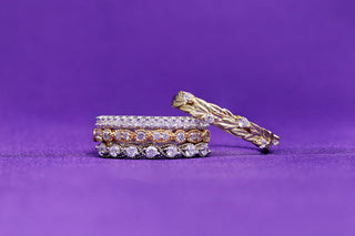 Stacked rose gold and silver pave eternity bands with a gold leaf-and-vine band leaning against them on purple fabric.