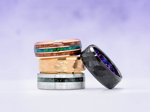 Four rings displayed together on a soft lavender background. The stack includes a black hammered ring leaning against the others, a silver-toned ring with a dark inlay, a gold-toned ring, and a rose gold-toned ring with a green and blue iridescent inlay.