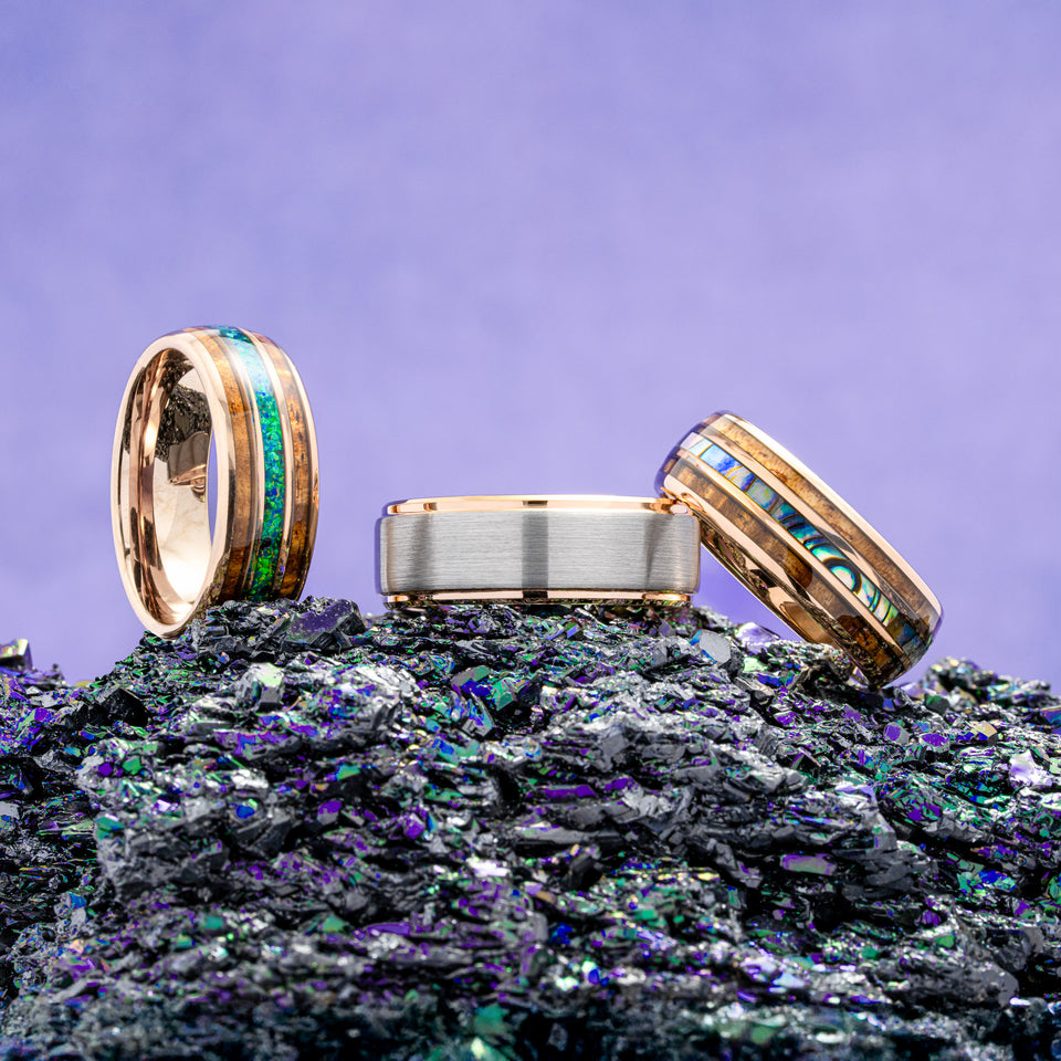 Three tungsten wedding bands with gemstone inlays on a textured surface with a purple gradient background