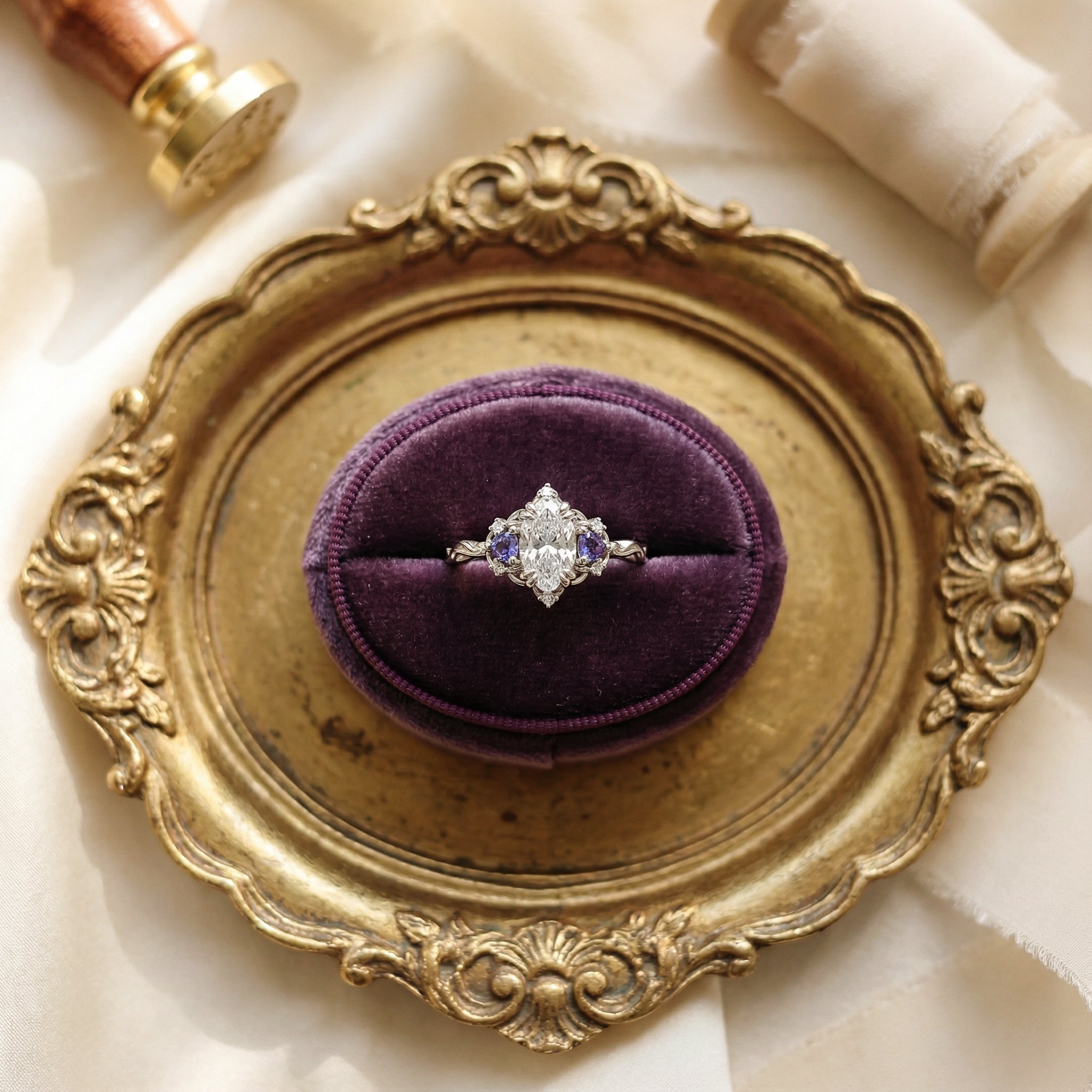 Vintage-style marquise diamond engagement ring with purple alexandrite side stones and an ornate white gold gallery, displayed in a purple velvet ring box on a gilded tray